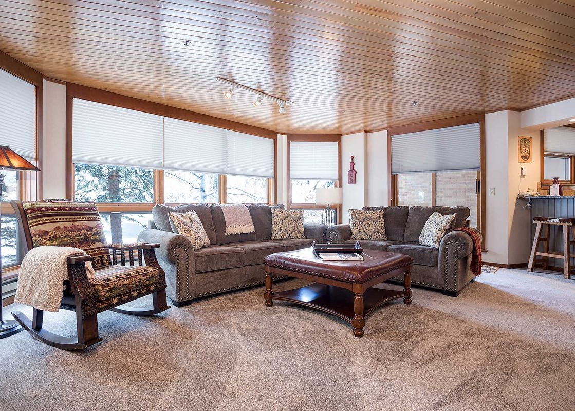 A cozy living room in Chateau Chamonix CX222 features a gray sofa, a rocking chair, a leather ottoman, and large windows offering a snowy outdoor view.