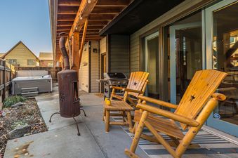 An inviting outdoor patio at Essentia Townhome 151B features a hot tub, BBQ grill, a rustic outdoor fireplace, and wooden Adirondack chairs, all set on a concrete slab with a surrounding privacy fence.