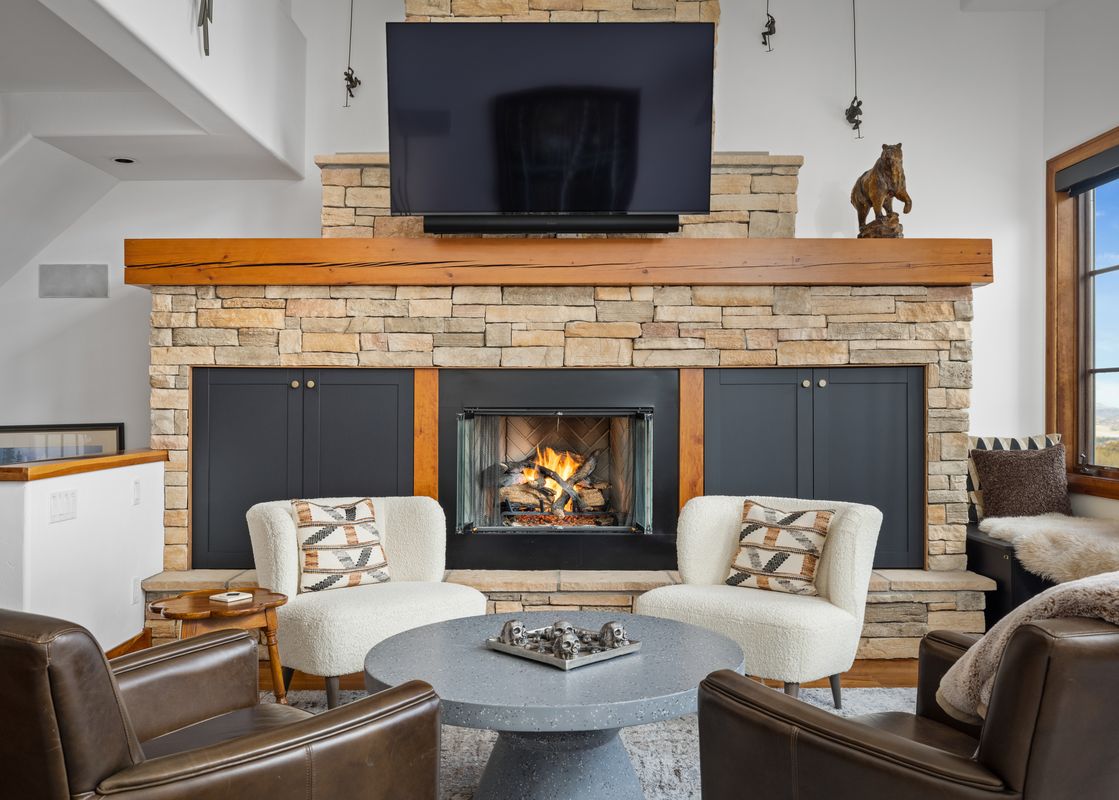 A cozy living room with a large stone fireplace, a flat-screen TV, and comfortable seating, offering a view of the mountains.