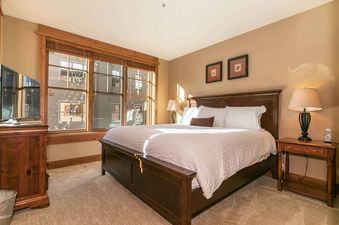 A bedroom with a large window, a king-sized bed with white linens, and a side table with a lamp.