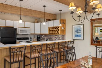 The image features the kitchen and dining area with white cabinets, a black refrigerator, a patterned tile backsplash, a breakfast bar with four stools, and a wooden dining table with wine, under a chandelier and exposed wooden beams.