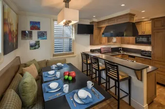 The image shows a dining area with bench seating and a set table, along with an adjacent kitchen featuring wooden cabinets and a breakfast bar.