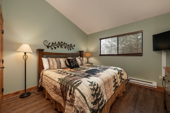 The bedroom at Beaver Village 333 features a queen-sized bed with a rustic-patterned comforter, flanked by a floor lamp and a bedside table with a lamp, all set against sage green walls.