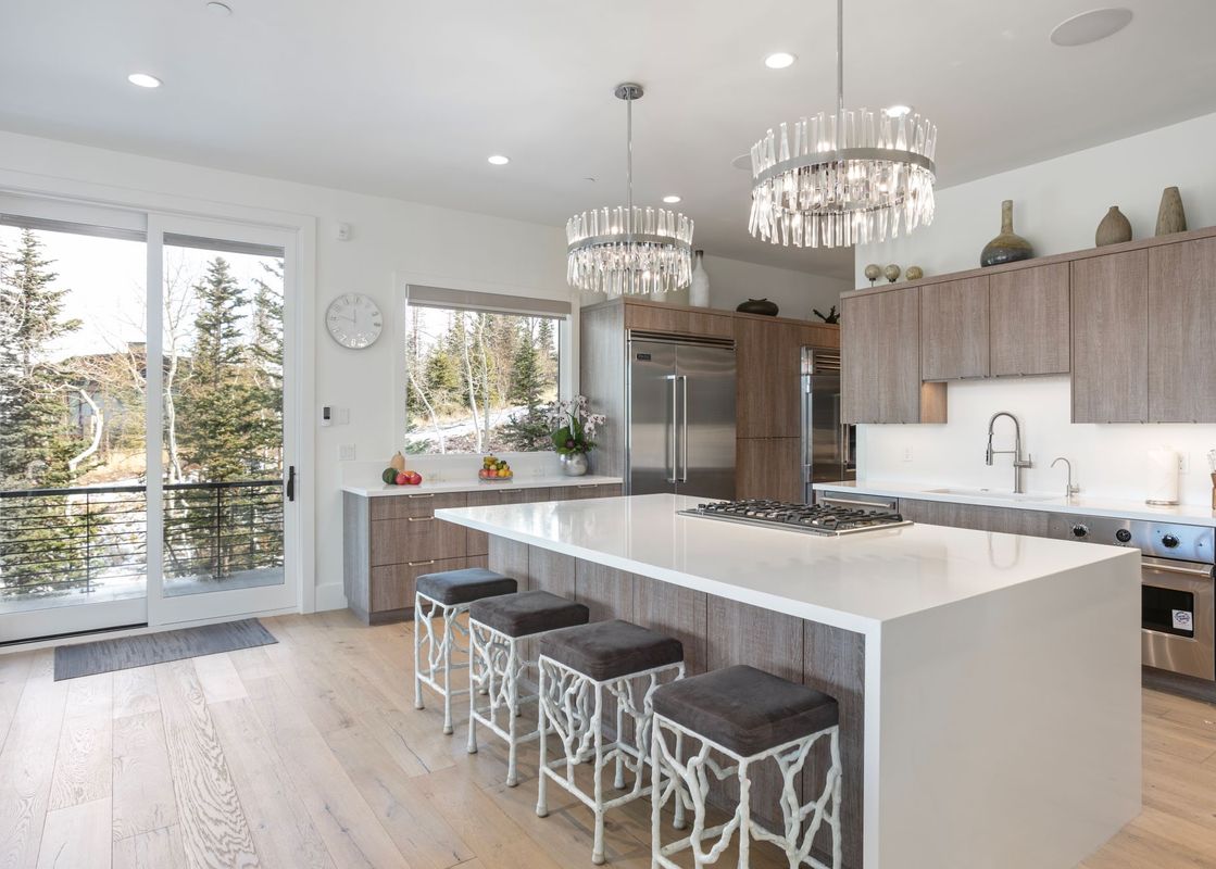 A modern kitchen with a large island, four bar stools, crystal chandeliers, stainless steel appliances, and a glass door leading to a balcony with a view of pine trees.