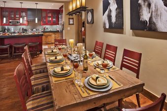 The dining area and kitchen of Sage Creek One Steamboat Place 105 are elegantly furnished, featuring a long wooden dining table set for a meal and a modern kitchen with red cabinetry.