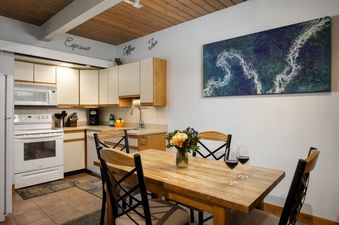 A fully equipped kitchen with white appliances and wood cabinets opens into a dining area with a wooden table, chairs, and a floral centerpiece, all beneath a unique wood-beamed ceiling.
