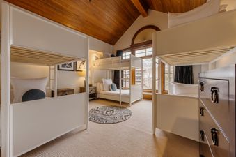 A cozy bunk room at High West at the Plaza features two sets of white bunk beds with white bedding, a light striped carpet, and a round patterned rug, all beneath a sloped wooden ceiling.