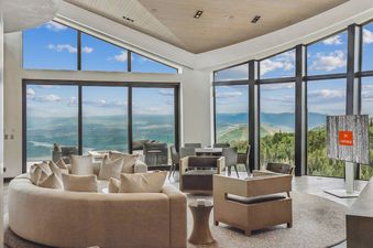 A luxurious and spacious lounge in Mountainhead with panoramic views of mountains and valleys through expansive windows, featuring a large, curved sectional sofa, contemporary armchairs, and a dining area.