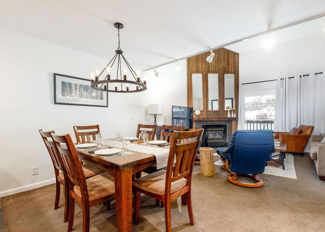 A wide view of the dining area and an adjacent living room at Snow Flower 129 Park City, featuring a wooden dining table and chairs, a fireplace, and comfortable seating.