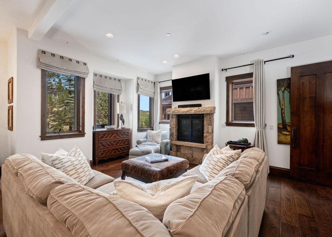 A cozy living room with a U-shaped sofa, stone fireplace, and views of nature from the windows.