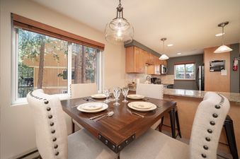A square wooden dining table is perfectly set for four, featuring classic round plates and elegant silverware. An inviting view of lush green trees can be seen through the large window.