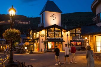 A charming European-style village square at dusk, with illuminated buildings, a clock tower, and people strolling, all framed by dark mountains in the background.