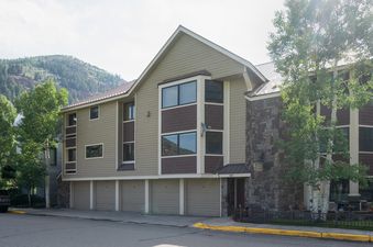 The exterior of The Sundance Condos 202, showing a multi-story building with beige siding, dark brown shingles, and stone accents, set against a backdrop of green mountains under a bright sky.