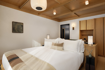 A modern and cozy bedroom features a large bed with white linens and a patterned throw, a warm wooden paneled ceiling, and contemporary furnishings.