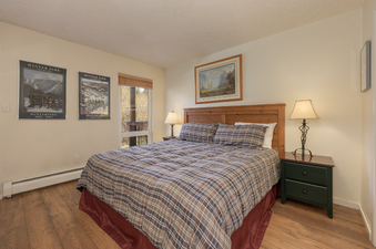 A cozy bedroom at Hi Country Haus 1803 features a comfortable bed with plaid bedding, wooden furniture, and framed artwork on the walls, creating a warm and inviting atmosphere.