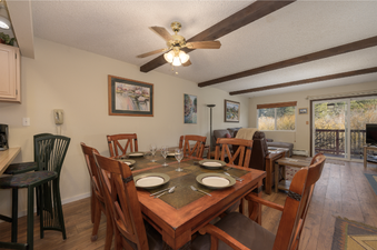 The interior of Hi Country Haus 1803 features a wooden dining table set for six, next to a living area with comfortable seating and a view of the outdoors through a large window and glass door.