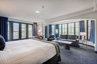 The Pines Lodge King Corner Room features a king-sized bed, a desk, a living area with a sofa and armchair, and large windows with blue drapery offering scenic views.