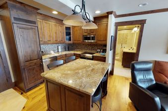 Kitchen with stainless steel appliances, granite countertops, and a breakfast bar with stools. The kitchen is open to a living room with a leather couch and a picture on the wall.