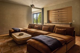 A luxurious and spacious lounge area with a large, plush sectional sofa, a unique coffee table, and a built-in wet bar, offering views of a serene natural landscape.