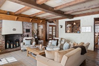 A rustic yet elegant living area at Five Trees Estate, featuring exposed wooden beams, a brick fireplace with lanterns, and comfortable floral armchairs and a beige sofa around a wooden coffee table.