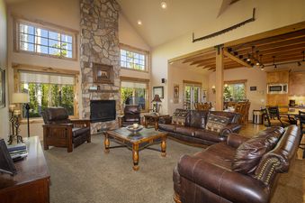 A spacious and inviting living room at Leland Creek Home, featuring comfortable leather sofas and armchairs, a large stone fireplace, and abundant natural light from numerous windows overlooking a lush, wooded exterior.