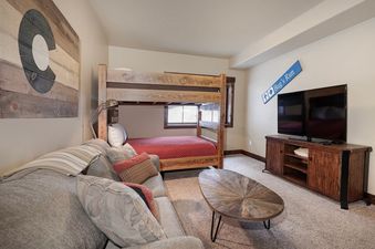 A cozy bedroom in 751 Mountain Terrace features a wooden bunk bed with a red comforter, a comfortable sofa, a rustic coffee table, and a TV on a wooden console, all set on a carpeted floor.