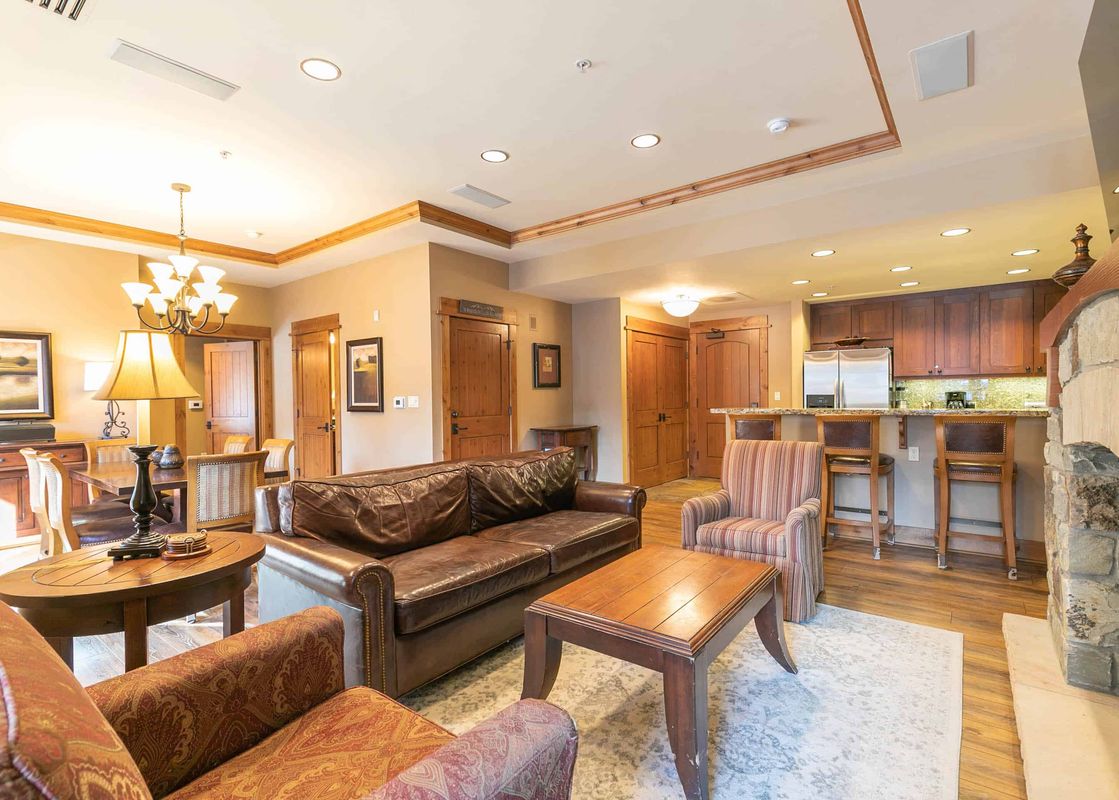 A living room with a leather couch, a coffee table and an armchair, a dining room to the left, and a kitchen and stone fireplace to the right.