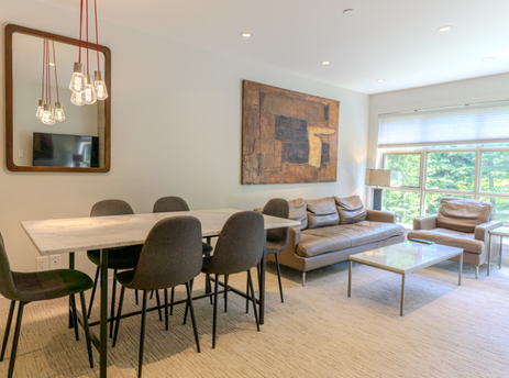 A modern and spacious living area is shown, featuring a dining table with chairs, a comfortable sofa, and large windows that offer a view of lush greenery outside.