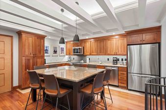 The kitchen features wooden cabinetry, granite countertops, stainless steel appliances, and a large island with bar stool seating under a white beamed ceiling with modern lighting.