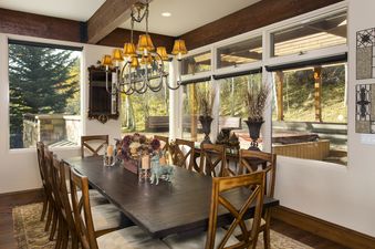 The dining area at 185 Nighthawk Dr features a large wooden table with several chairs, an ornate chandelier, and expansive windows offering views of a forested landscape and an outdoor hot tub.