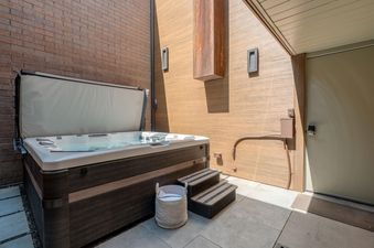 A private outdoor hot tub with a partially open cover, wooden steps, and a basket of towels, set against brick and wood-paneled walls.