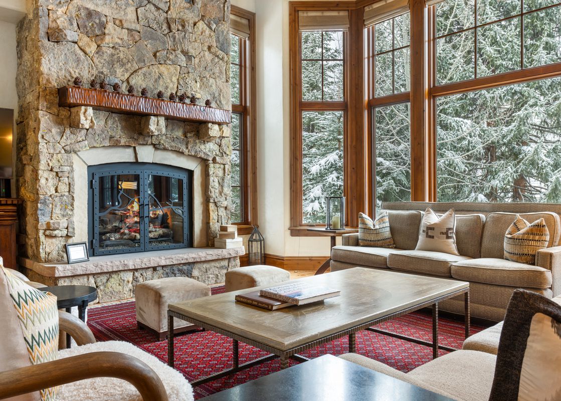 A living room with a large stone fireplace, a couch, and a coffee table, with large windows looking out onto a snowy forest.