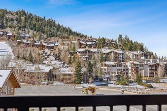 An expansive outdoor shot captures the snow-dusted roofs of numerous brown and white buildings nestled amidst a dense forest, with a clear blue sky overhead and a snowy ski path winding down the mountainside.
