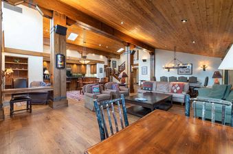 A spacious, open-plan great room with rustic wooden beams, plush seating, and a dining area, bathed in warm lighting.