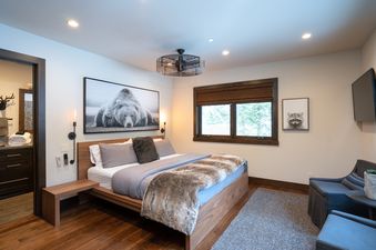 The image shows the master bedroom at West Shore Hideaway, featuring a king-sized bed, a large window with a Roman blind, a mounted television, and a cozy seating area.