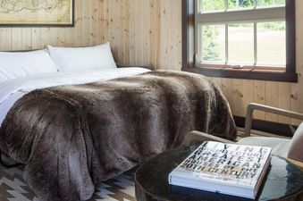 A cozy bedroom in The Shooting Cabin featuring a bed with a faux fur throw, a gray sofa, and a window overlooking a green landscape.