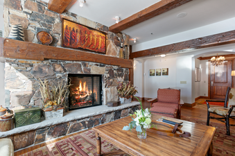 A cozy living area in Grand Lodge Three Bedroom Deluxe 404, showcasing a stone fireplace with a roaring fire, wooden beams, and large windows offering a view of the surrounding mountains.
