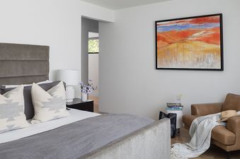 A modern bedroom with a gray upholstered bed, a vibrant abstract painting on the wall, and a cozy brown leather armchair.