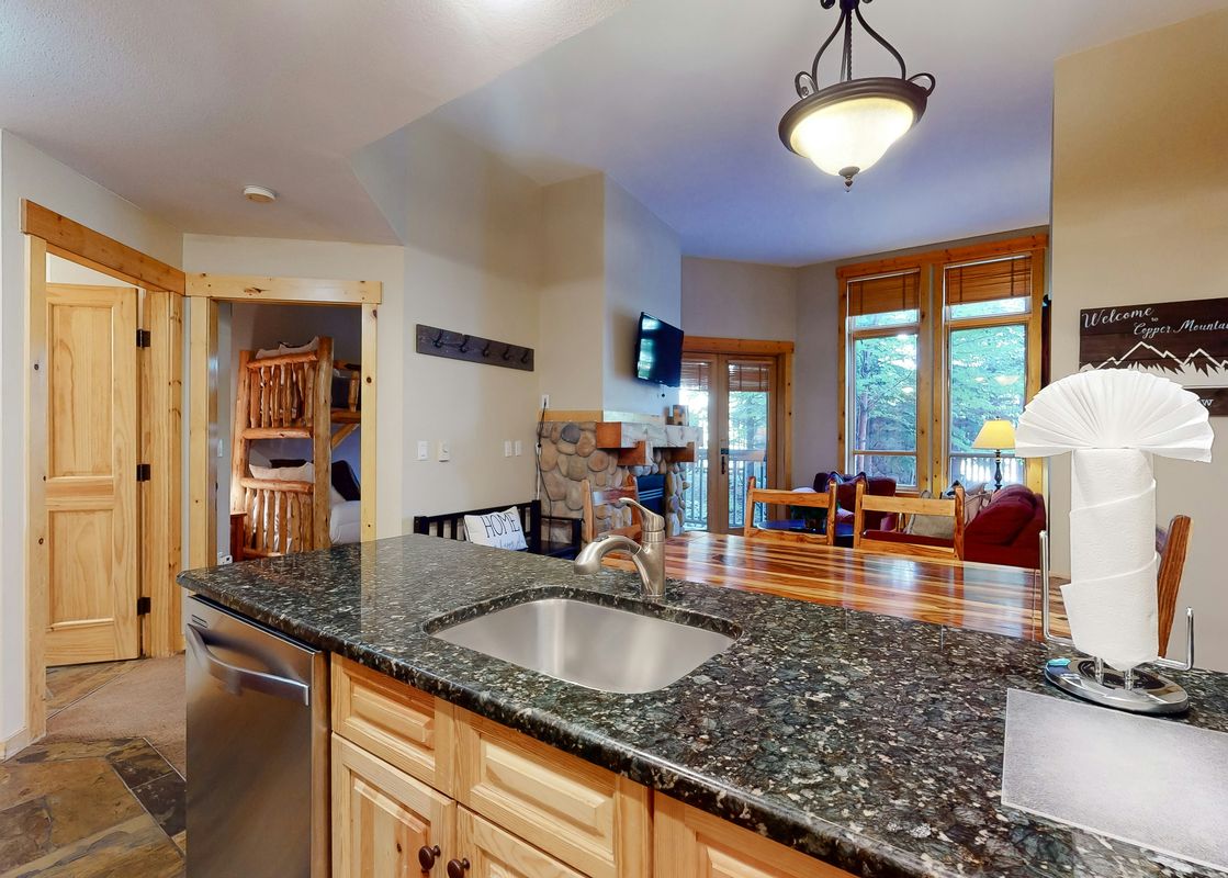 The kitchen at Taylors Crossing 109 features dark granite countertops, a stainless steel sink, and light wood cabinetry, with a cozy living area visible in the background.