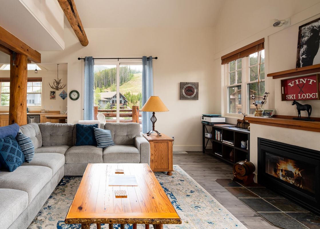 A spacious living room features a comfortable sectional sofa, a large coffee table, a fireplace, and a window with a view of the mountains.