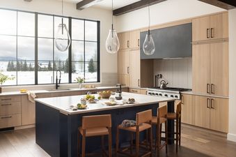 A modern kitchen with light wood cabinets, a dark blue island, leather bar stools, and large windows revealing a snowy mountain landscape.
