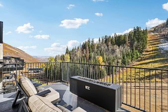 The balcony at Founders Place 2502 offers comfortable seating and a fire pit, overlooking a scenic mountain landscape with autumn foliage and ski slopes under a blue sky.