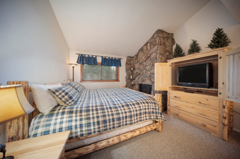 A cozy bedroom in Beaver Village 1134 features a rustic wood bed frame with plaid bedding, a stone fireplace, and a light wooden entertainment cabinet with a television.