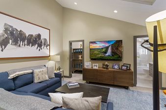 A living room with a blue sectional sofa, a coffee table, a large flat screen television, a wooden cabinet, and a bathroom with a shower and toilet visible in the background.