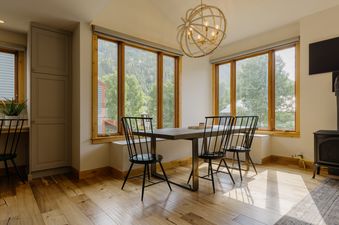 The dining area at Telluride Lodge Retreat features a large wooden table with black chairs, surrounded by windows that offer views of the lush green mountainside.