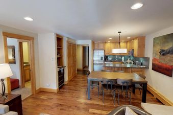 A spacious living area with a dining table and chairs in the center, a fully equipped kitchen on the right, and an open doorway leading to another room in the background.