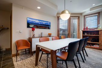 A modern dining area with a white-topped table, tan and black chairs, and a large mountain landscape artwork on the wall.