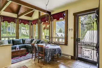 The image shows the open-concept living and dining area of the Summer Haus, featuring a dark green sectional sofa, a dining table with a blue floral tablecloth, and large windows offering views of the outdoors. Wooden exposed beams add to the rustic charm