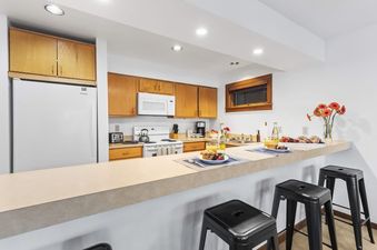 A fully equipped kitchen with wooden cabinetry, white appliances, and a breakfast bar with black stools.
