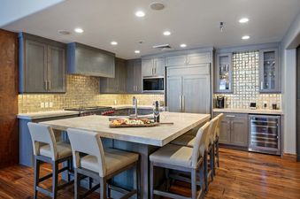 The modern kitchen in Grand Lodge 5 Bedroom Penthouse 504 boasts a large island with seating for guests. The cabinets are a dark grey wood, and the counters are a light grey stone. There are stainless steel appliances, including a refrigerator, microwave,